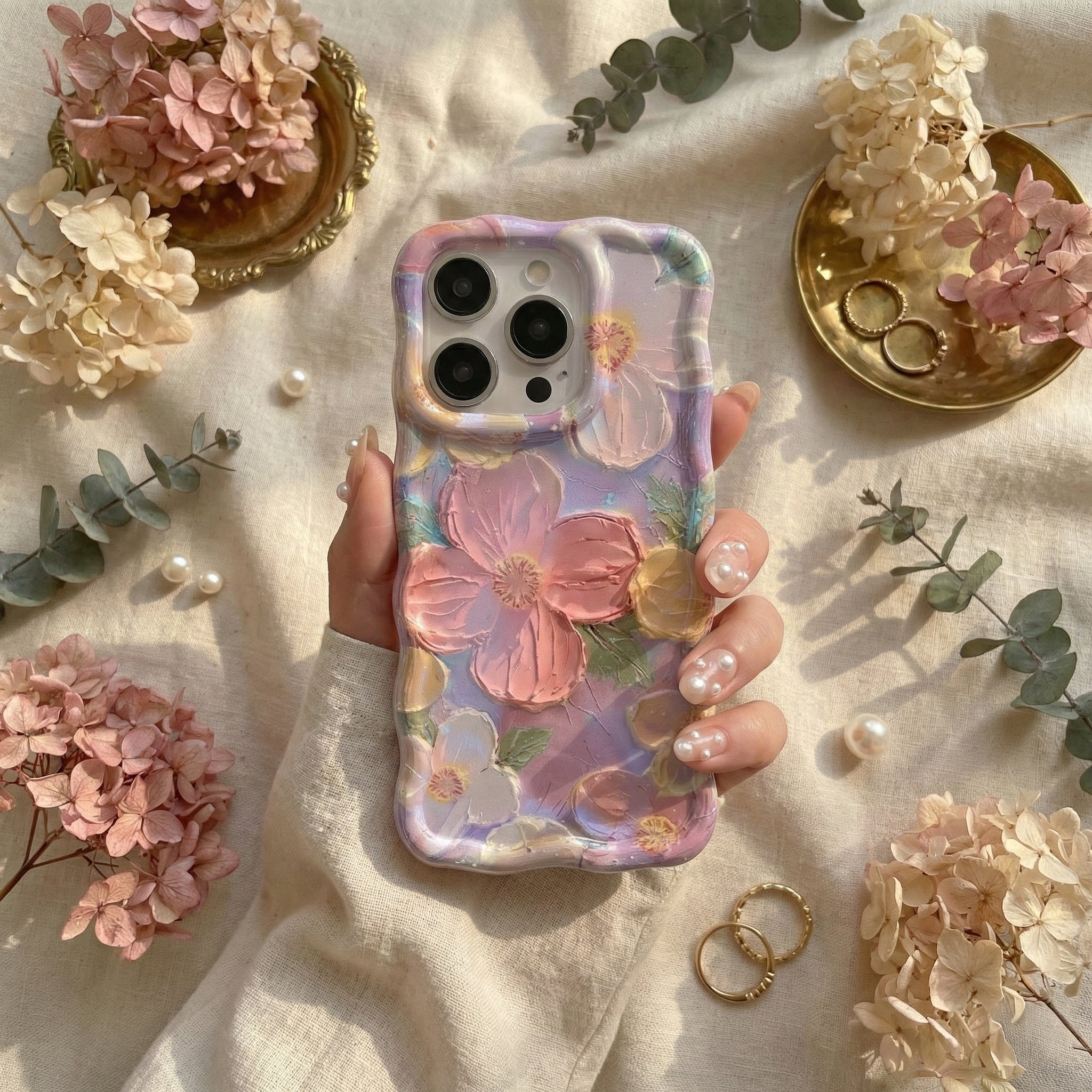 Phone case with floral design held by a hand, surrounded by flowers and jewelry on a light fabric background